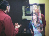 A color image of a man and a woman standing facing each other. The man in the red shirt, with his back facing the viewers, points to himself; the woman in the blue top puts her hands on her hips and stares back at the man. A reflection of the man's irate facial expression is cast on a glass surface, overlapping onto the woman's body.
