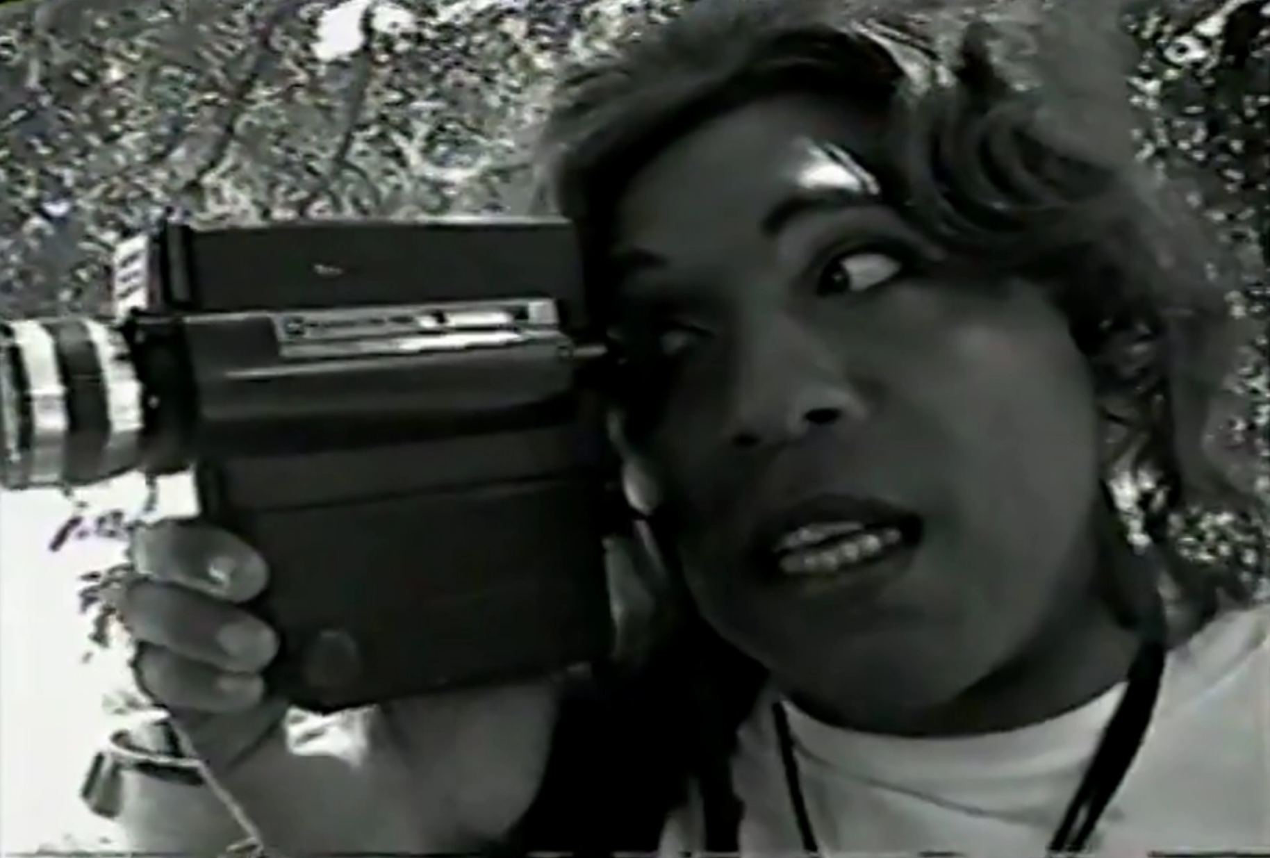 A grainy black and white photo of a woman craning her head and shoulders, looking into a large camera. In the background there are trees with leaves.