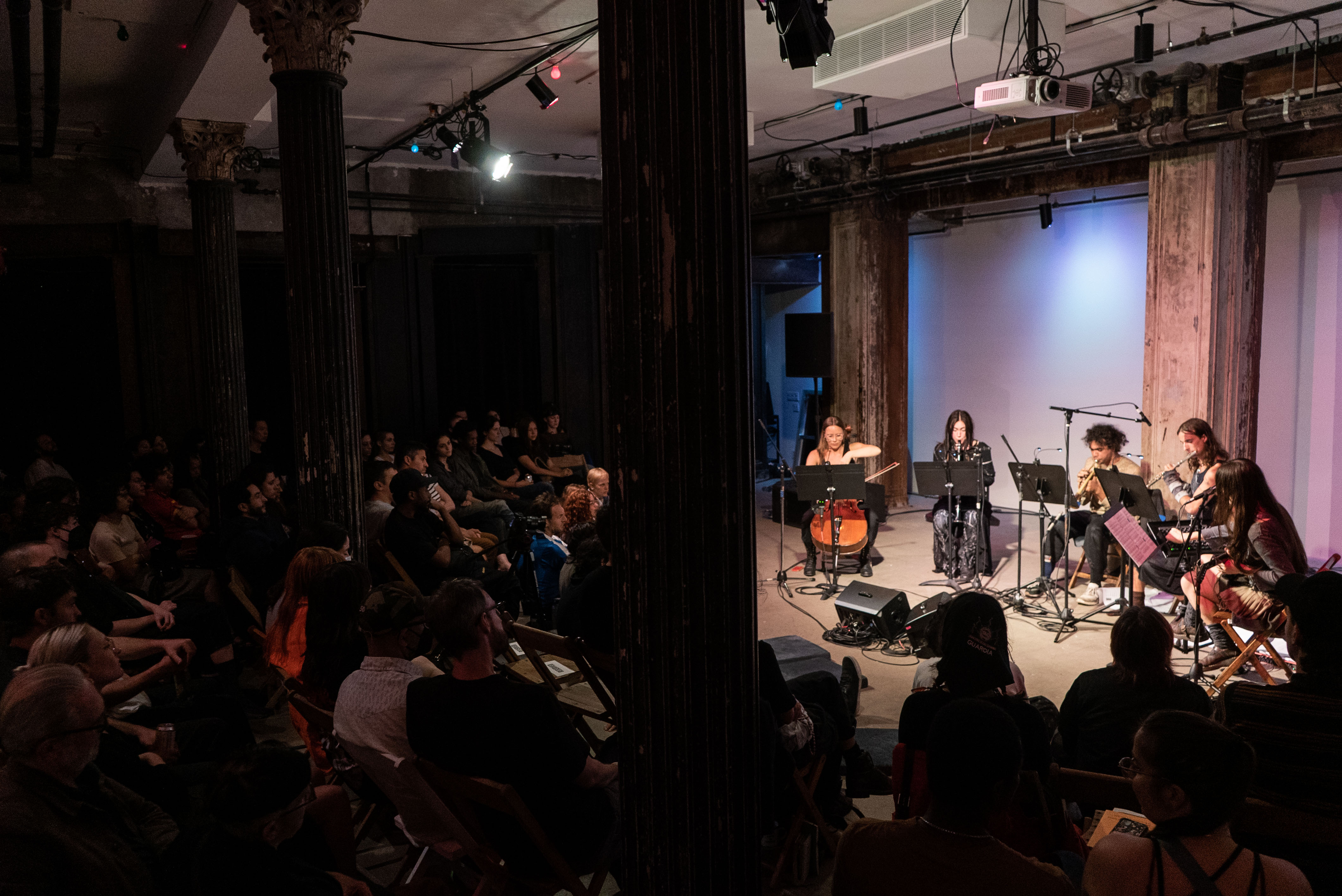 Wide view of 5 musicians on stage performing under blue tinted lights with the audience on the left. From left to right the instruments played are the cello, clarinet, horn, flute, guitar, and vocals.