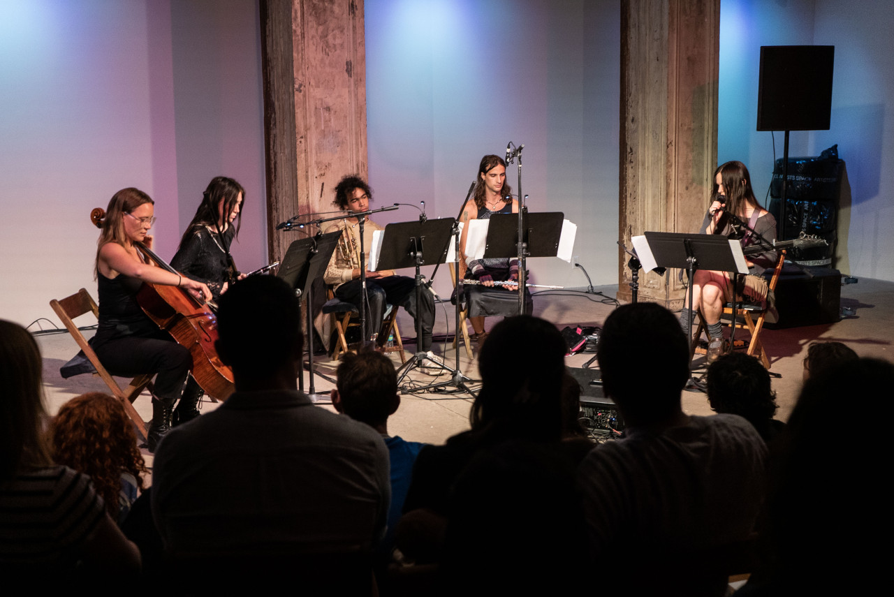 View of 5 musicians on stage performing under blue tinted lights with the audience in the foreground. From left to right the instruments played are the cello, clarinet, horn, flute, guitar, and vocals.