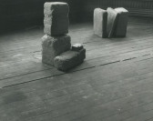 Installation view of several cast concrete blocks arranged on top of one another and leaning against each other, in two groups, standing on a wood floor.