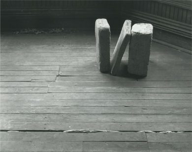 Installation view of three cast concrete blocks - the middle one leaning against the last one - standing on a wood floor.