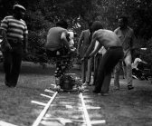 A black-and-white photograph of a group of men shooting a scene on an outdoor film set. They are surrounded by trees and grass.