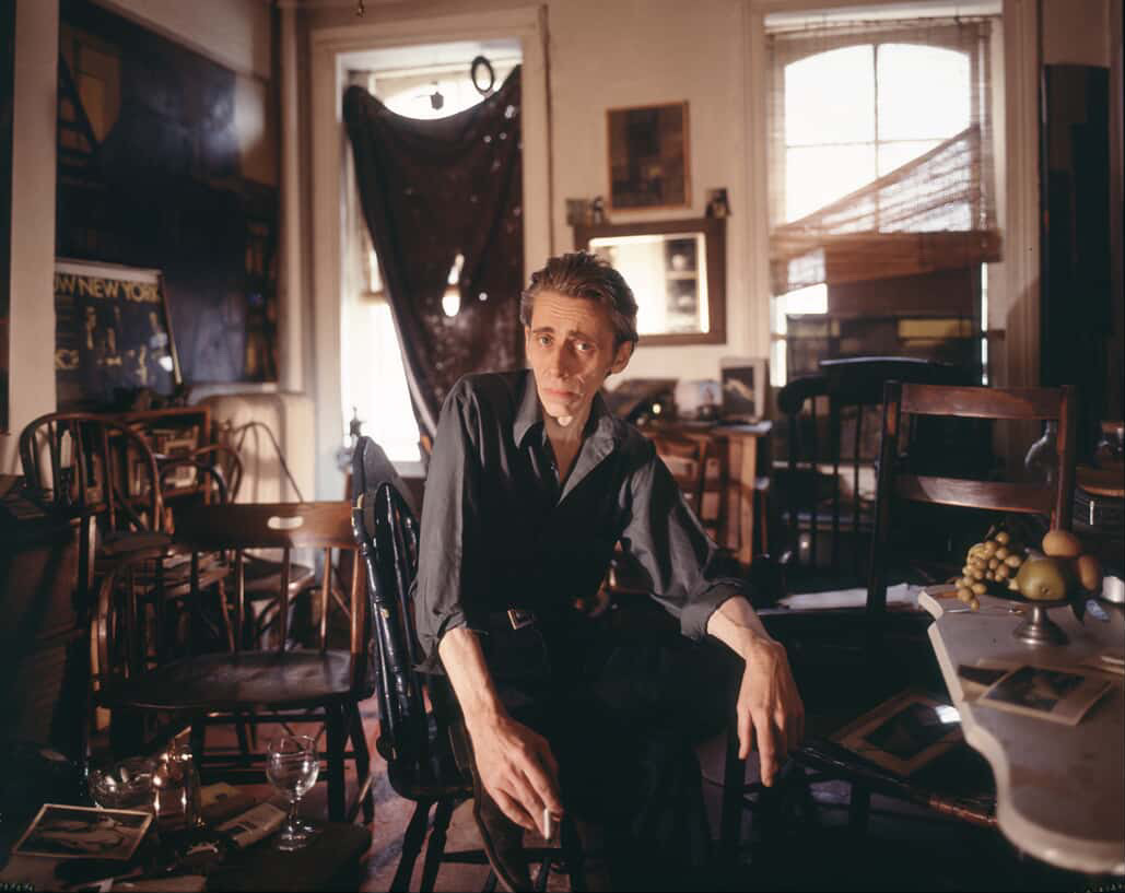 A male figure sits inside a crowded room with lots of wooden chairs, and large paintings and posters hung on the walls. Light streams in from the windows in the back.