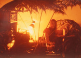 An orange-tinted image of a woman partially obscured by a table full of electronic equipment and a large plant.