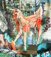 A young deer made out of puzzle pieces with red streamers coming out of its eye and hip, as if bleeding. It is on a multimedia background showing bricks and grass.