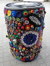 A color image of a decorated waste container on the street. The container is adorned with bottle caps in different colors, sizes, and materials, nailed to the surface of the bin. The dense arrangement of the caps on its surface creates complex color patterns and textural accentuations.
