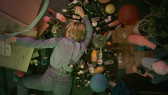 Color image of a white, blonde woman laying face down on the floor. Empty bottles of alcohol and other drinks cover the floor, along with balloons, cardboard boxes and a wooden shelf.