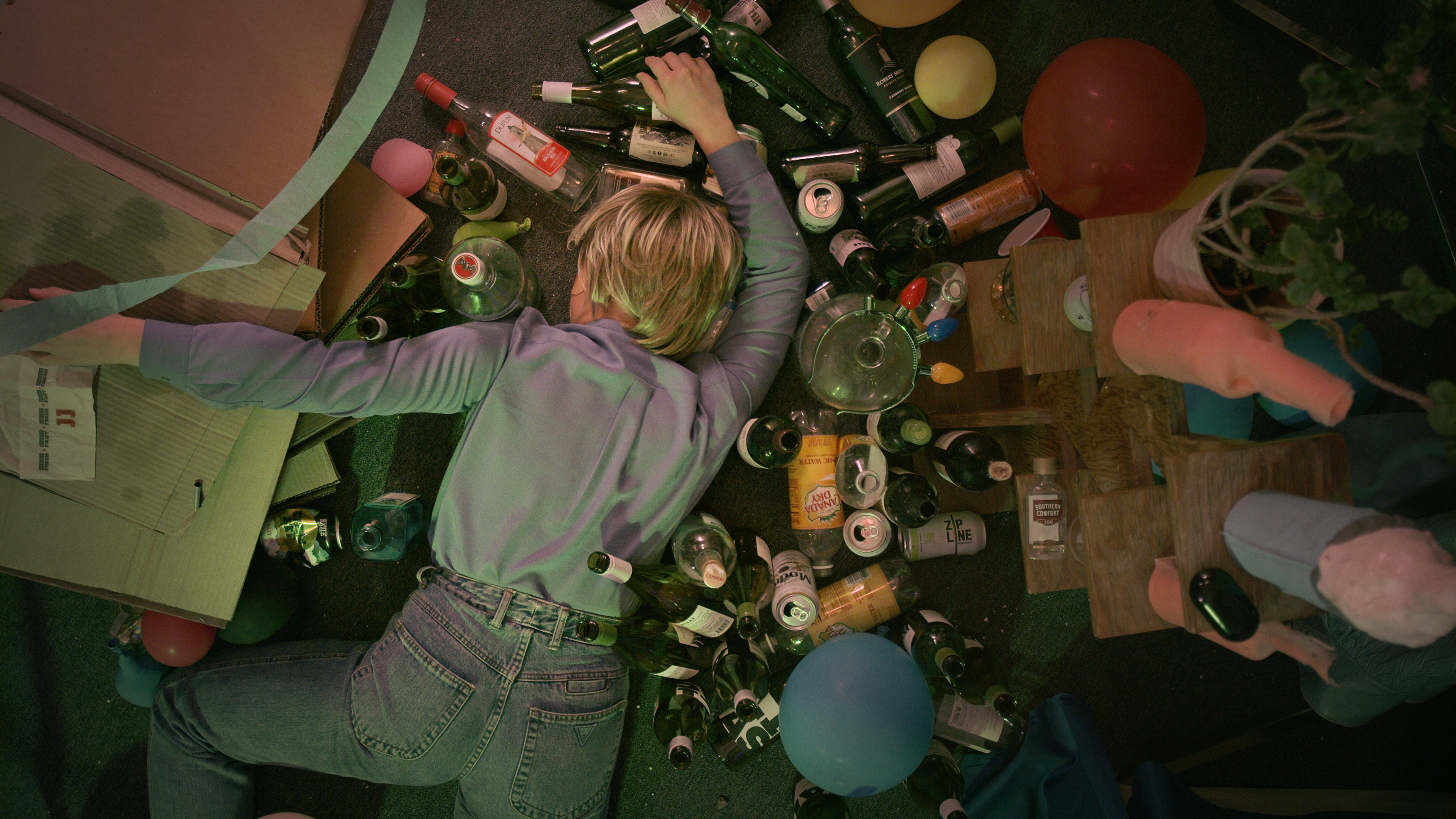 Color image of a white, blonde woman laying face down on the floor. Empty bottles of alcohol and other drinks cover the floor, along with balloons, cardboard boxes and a wooden shelf.