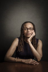 Portrait of a black woman, looking forward with her face resting in her left hand, and her right arm placed on a table. Behind her is a dark grey background.