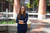 A black woman stands, smiling, with her arms crossed in front of her. Behind her is a small fountain inside a courtyard.