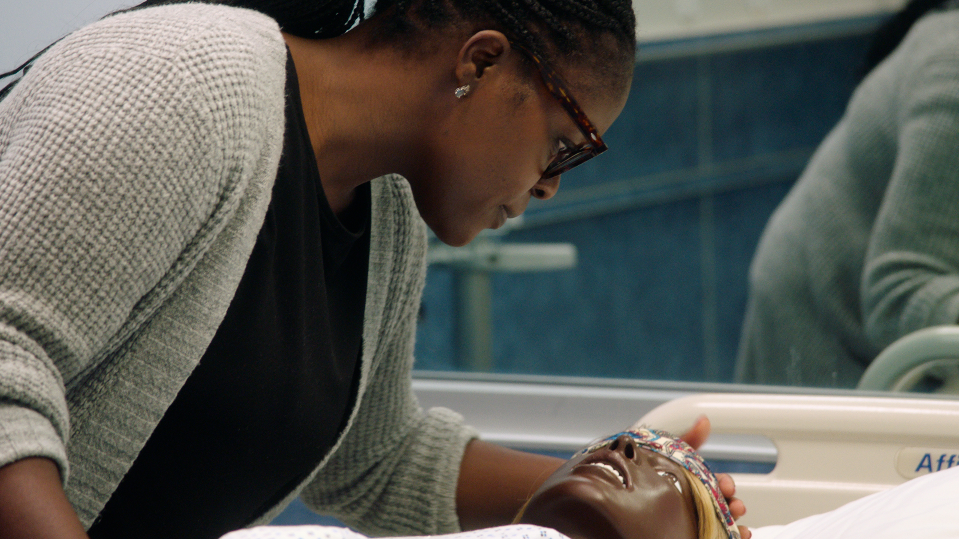 Close-up of a black person leaning over a black birthing mannequin laying on a hospital bed in a patient room.