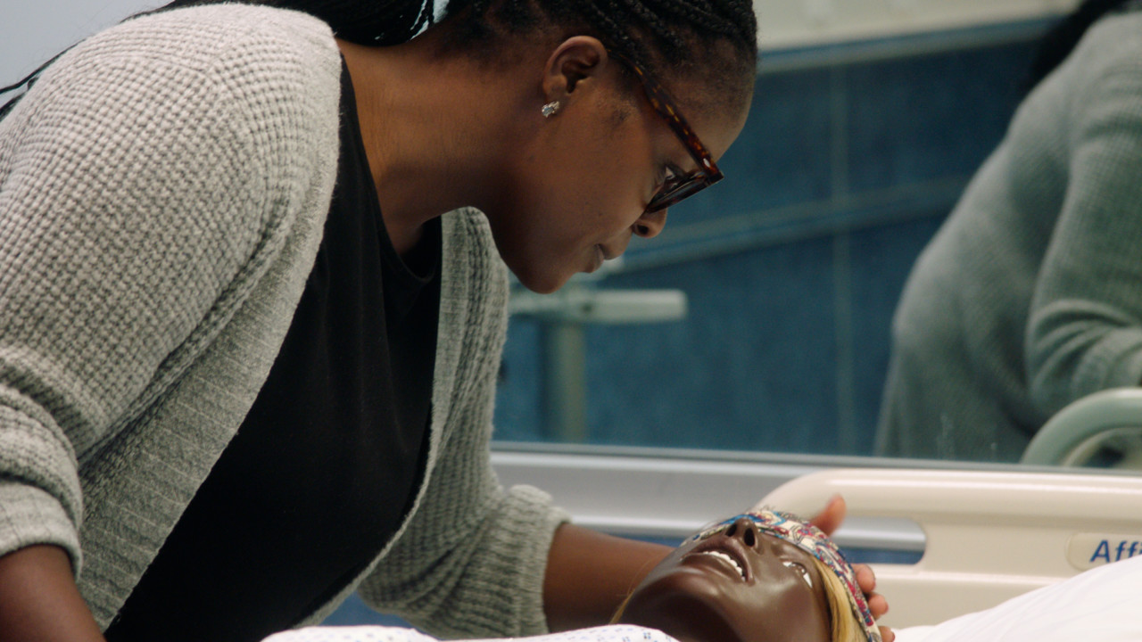 Close-up of a black person leaning over a black birthing mannequin laying on a hospital bed in a patient room.