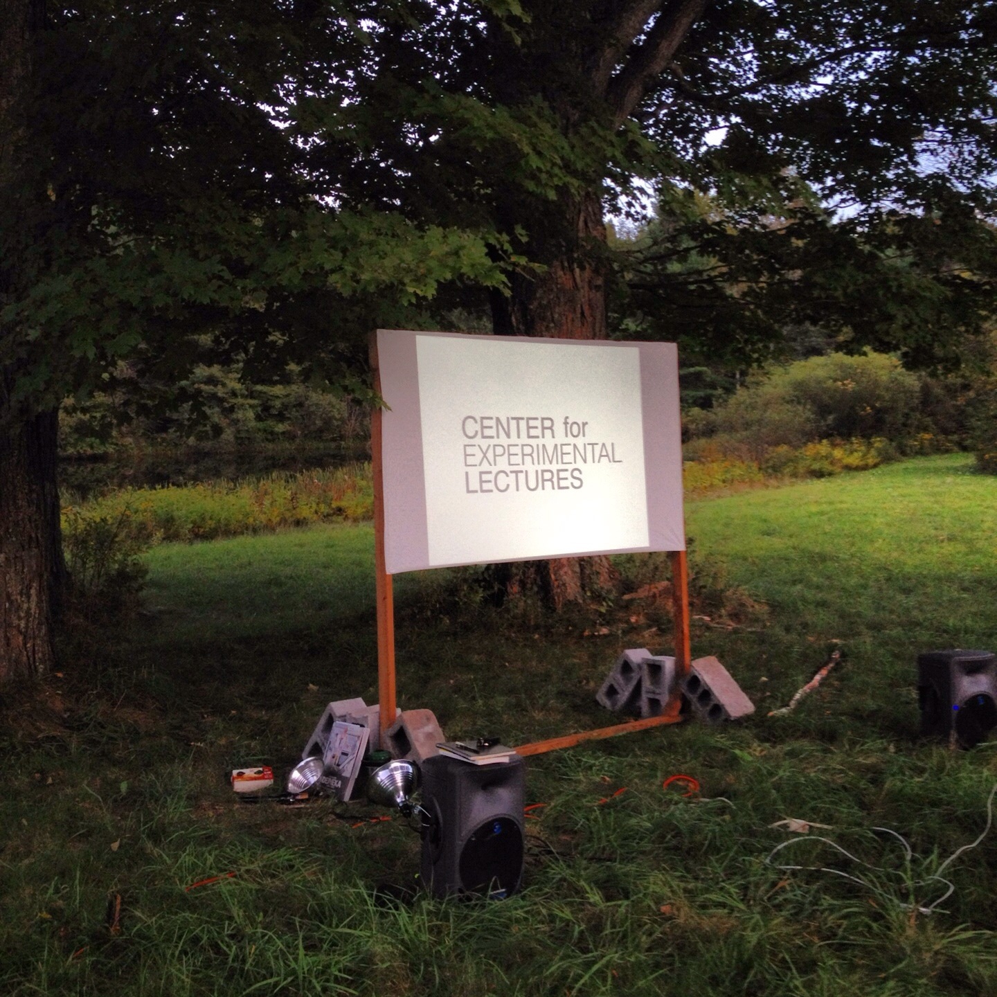 In a grassy clearing in the shade of two green oak trees mostly obscuring the sky, the words “CENTER for EXPERIMENTAL LECTURES” are projected onto a small screen supported by a wooden frame held upright between leaning cinderblocks. Small monitor speakers are in the foreground on either side, one with a clamp light attached to it and a stack of books on top of it.