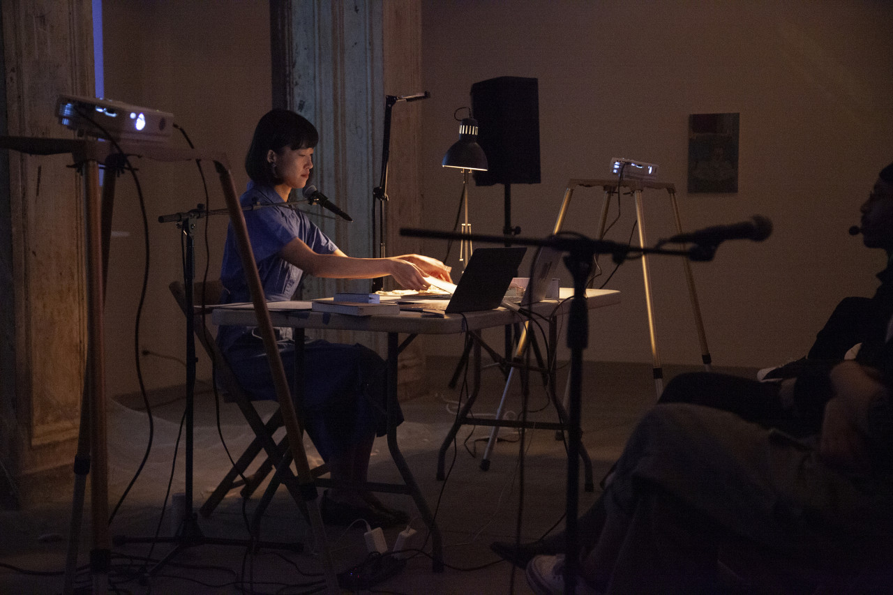 A figure in a blue dress sits behind a white table, with speakers and projectors on either side of them. The figure leans over and opens a small book.