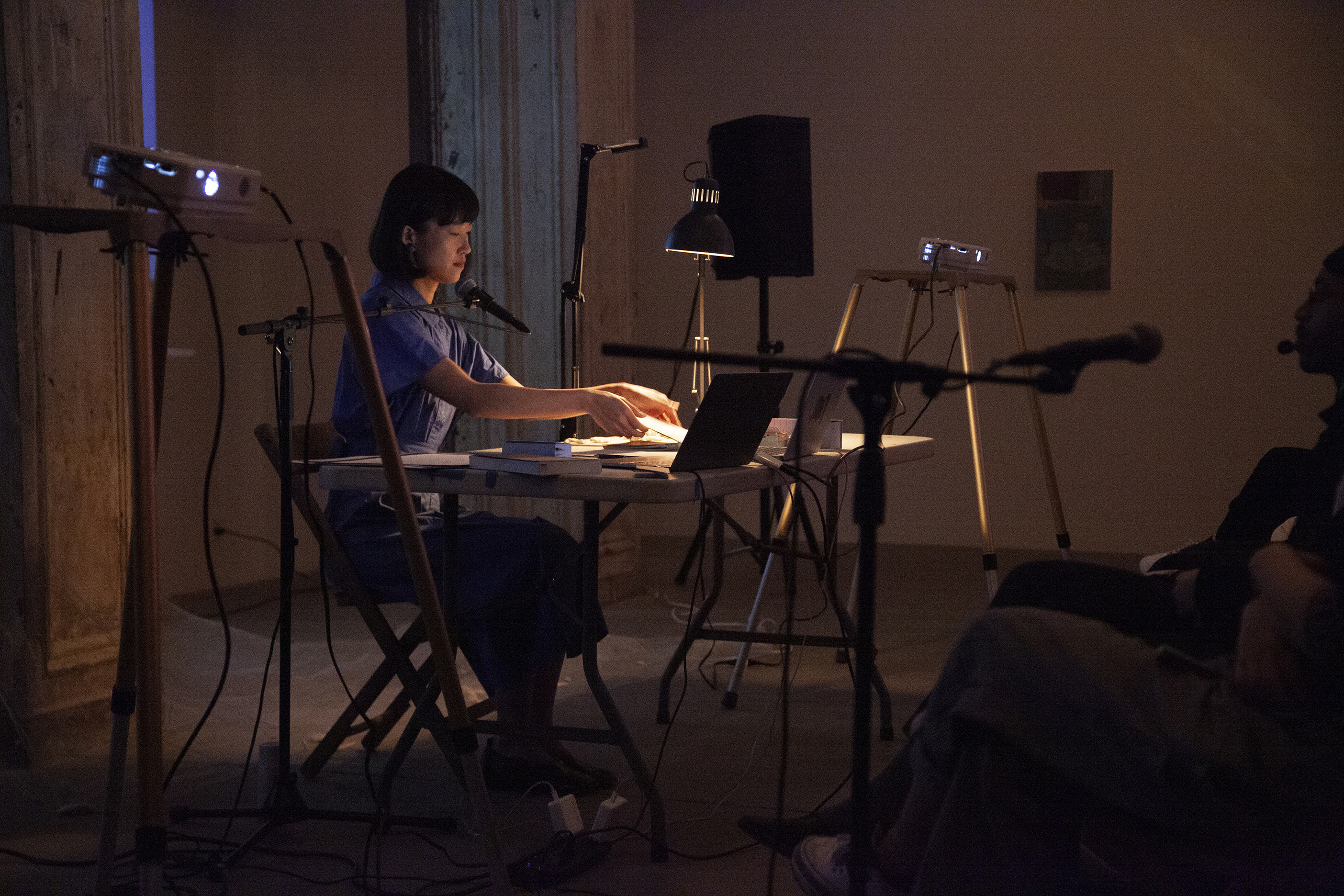 A figure in a blue dress sits behind a white table, with speakers and projectors on either side of them. The figure leans over and opens a small book.