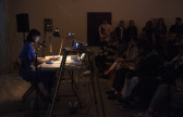 A figure in a blue dress sits behind a white table, they are reading from a small book in front of them. Projectors on either side of the table face the back wall. An audience watches the reader from the right side of the image.