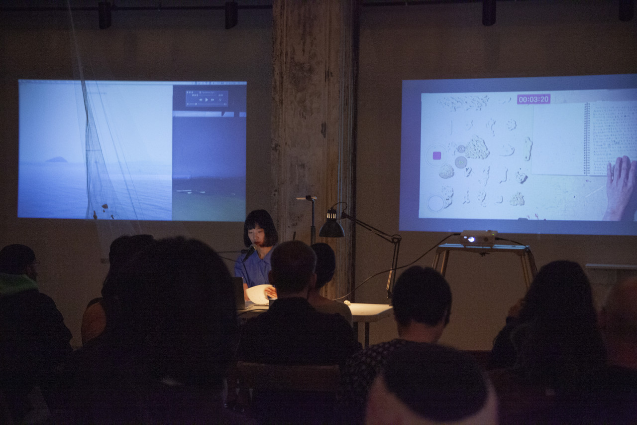 A figure in a blue dress sits behind a white table, facing an audeince and reading into a microphone. On the wall behind the presenter are two projected screens. The right screen depicts a table with a notebook, someone