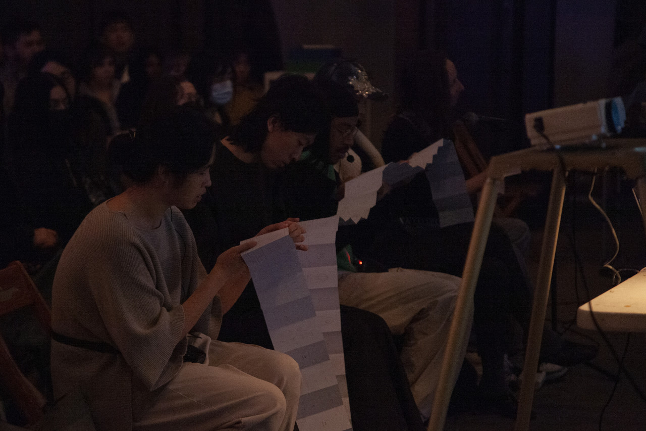 A row of figures sitting in wooden chairs hold various sections of a folded up book, reading off different parts.
