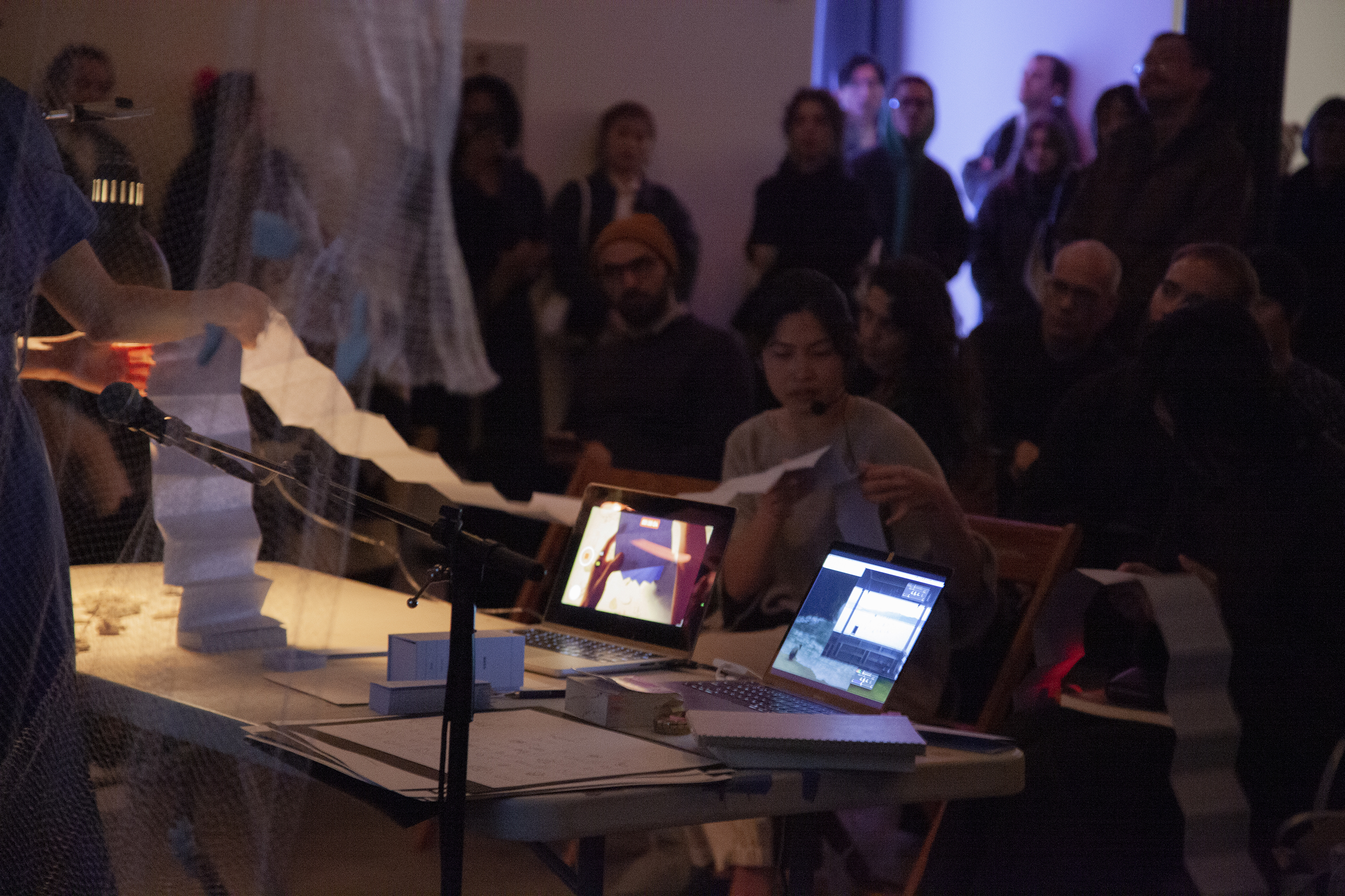 A figure's hands are shown, standing at the edge of a white table, unfolding a small book as another figure on the other side of the table holds part of the book and reads off of it.