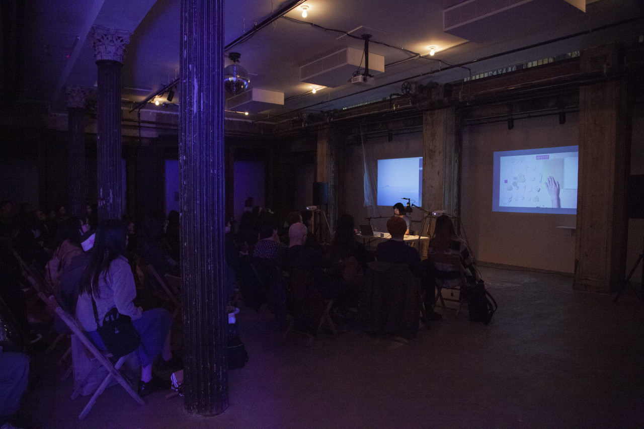 In a basement space, an audience watches as a figure sitting behind a white table reads into a microphone. On the back wall behind the figure are two projected videos featuring a notebook, pieces of coral, and an image of the ocean.