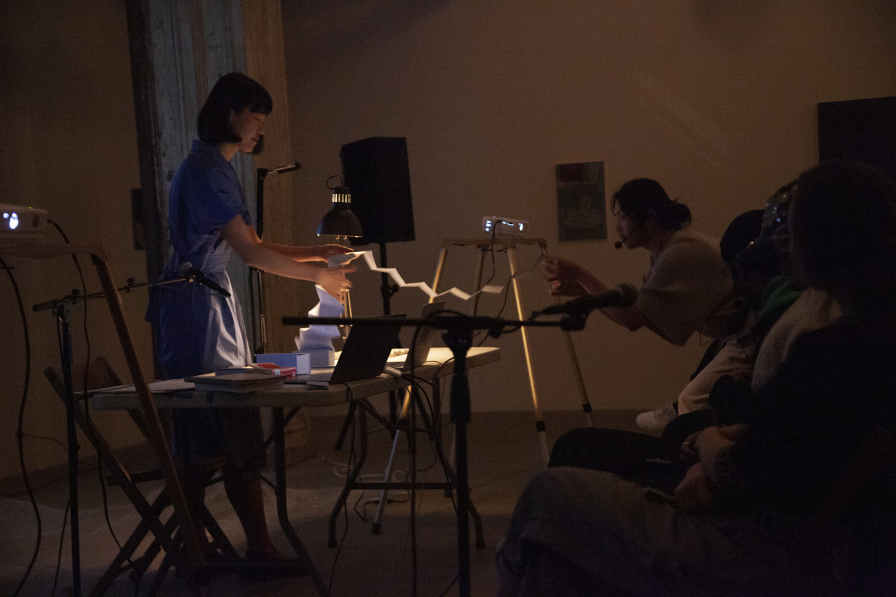 A figure in a blue dress stands behind a white table, they slowly unfold a book as another figure in the audience stands and reaches over to read off of the paper.