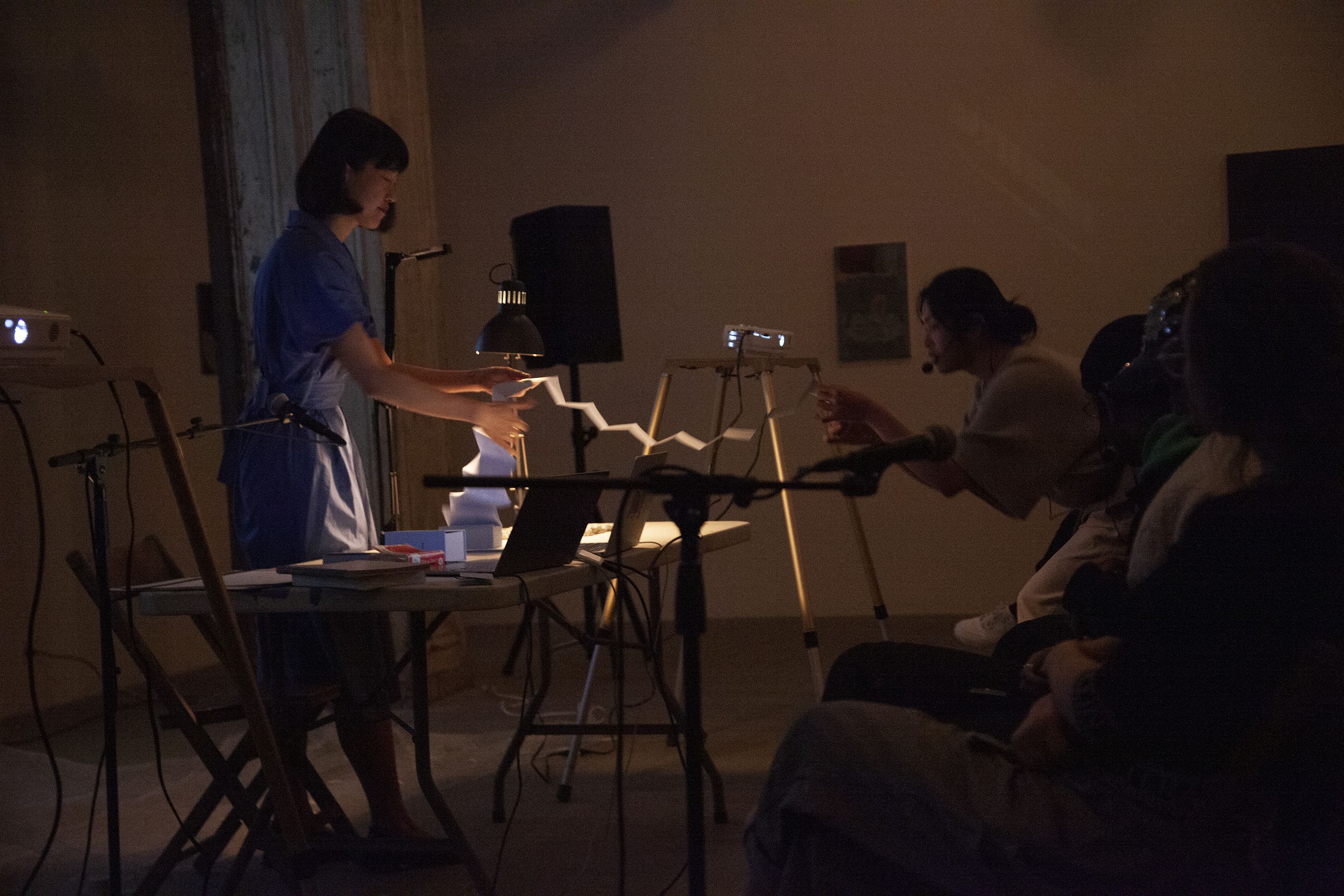 A figure in a blue dress stands behind a white table, they slowly unfold a book as another figure in the audience stands and reaches over to read off of the paper.
