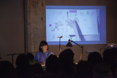 figure in a blue dress sits behind a white table, facing an audience. On the wall behind the presenter is a projected video that depicts an array of dried coral pieces on the left, and next to them a hand places a paper with illustrations of coral and a chart with notes underneath.