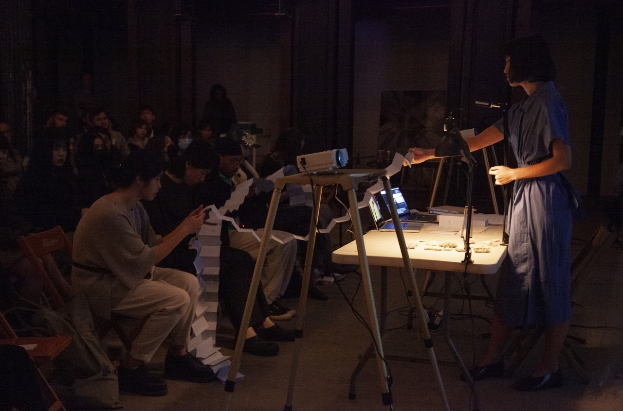 A figure in a blue dress stands behind a white table, and holds the end of a small folded up book. On the other side of the table, a row of people sitting on wooden chairs hold different sections of the book and read off of it.