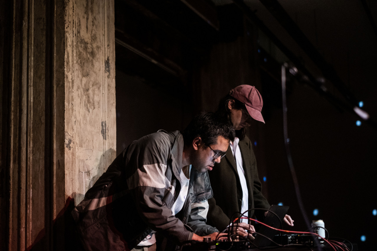 Two figures stand on stage, performing with audio equipment on a table in front of them.
