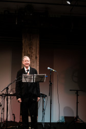 A figure in a suit stands on stage, behind a music stand and two microphones.