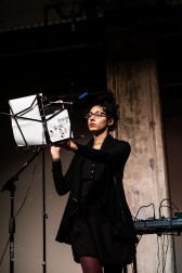A figure performs on stage behind a music stand.