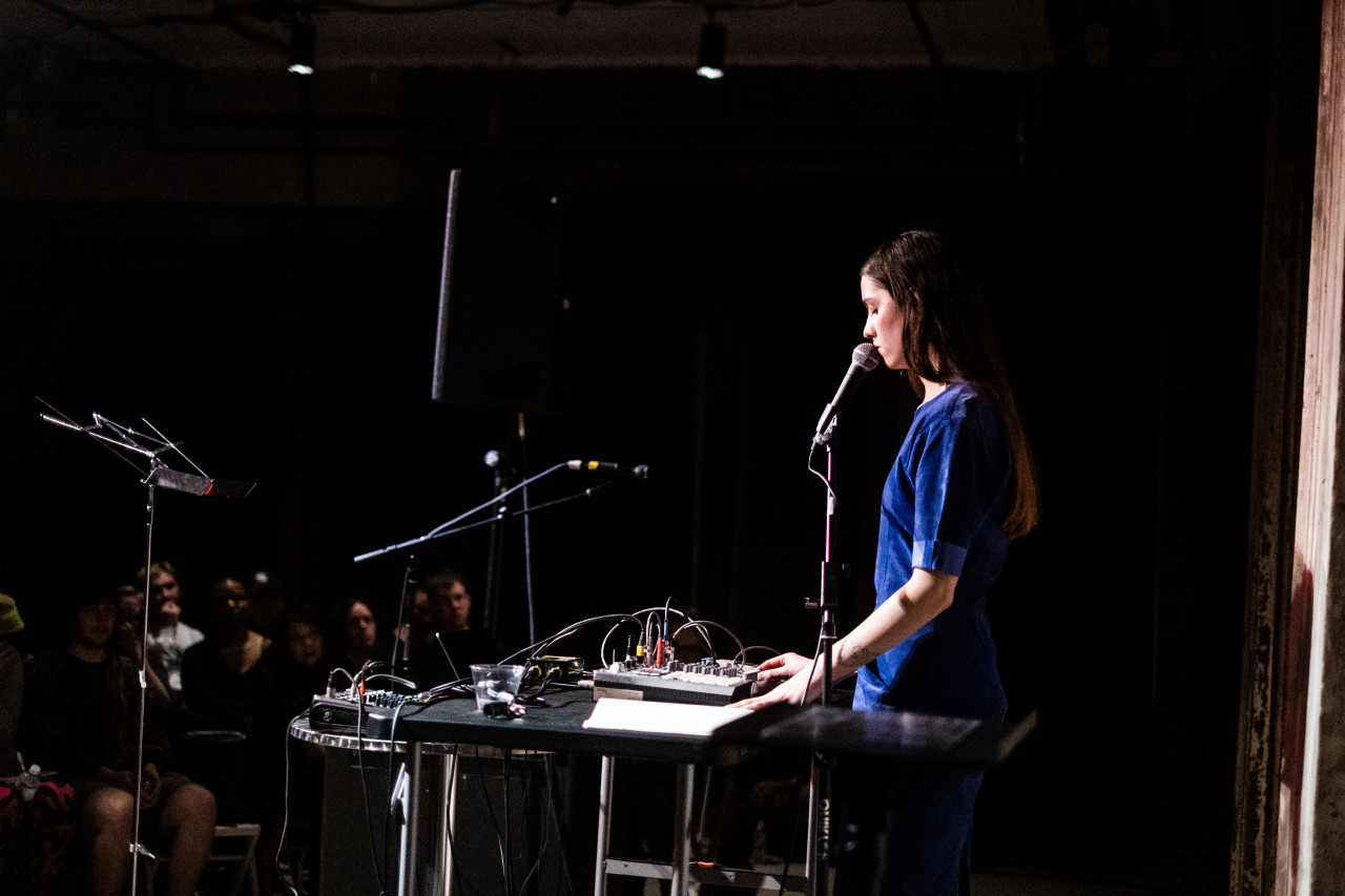 A figure performs on stage using a microphone and an audio mixer, with the audience in the back left of the image.