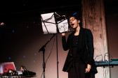 A figure performs into a microphone, their mouth open, performing using material held on a music stand in front of them.