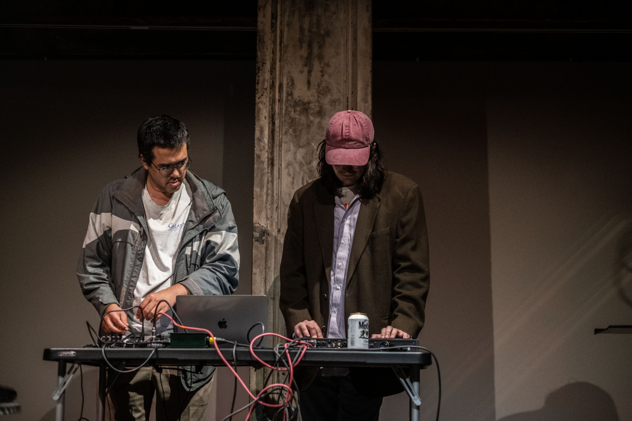 Two figures stand behind a table, setting up audio equipment.