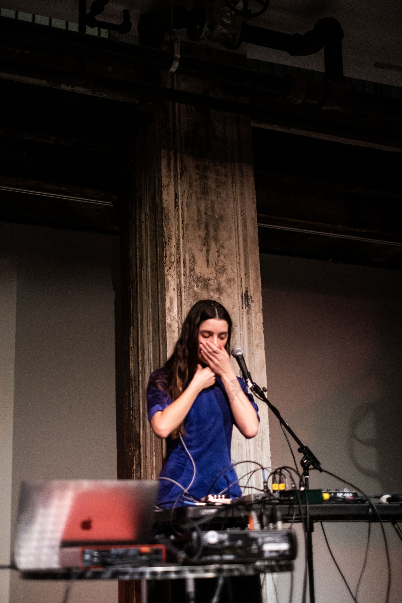 A figure stands behind a microphone and table with audio equipment. They hold one hand over their mouth and the other around their neck.