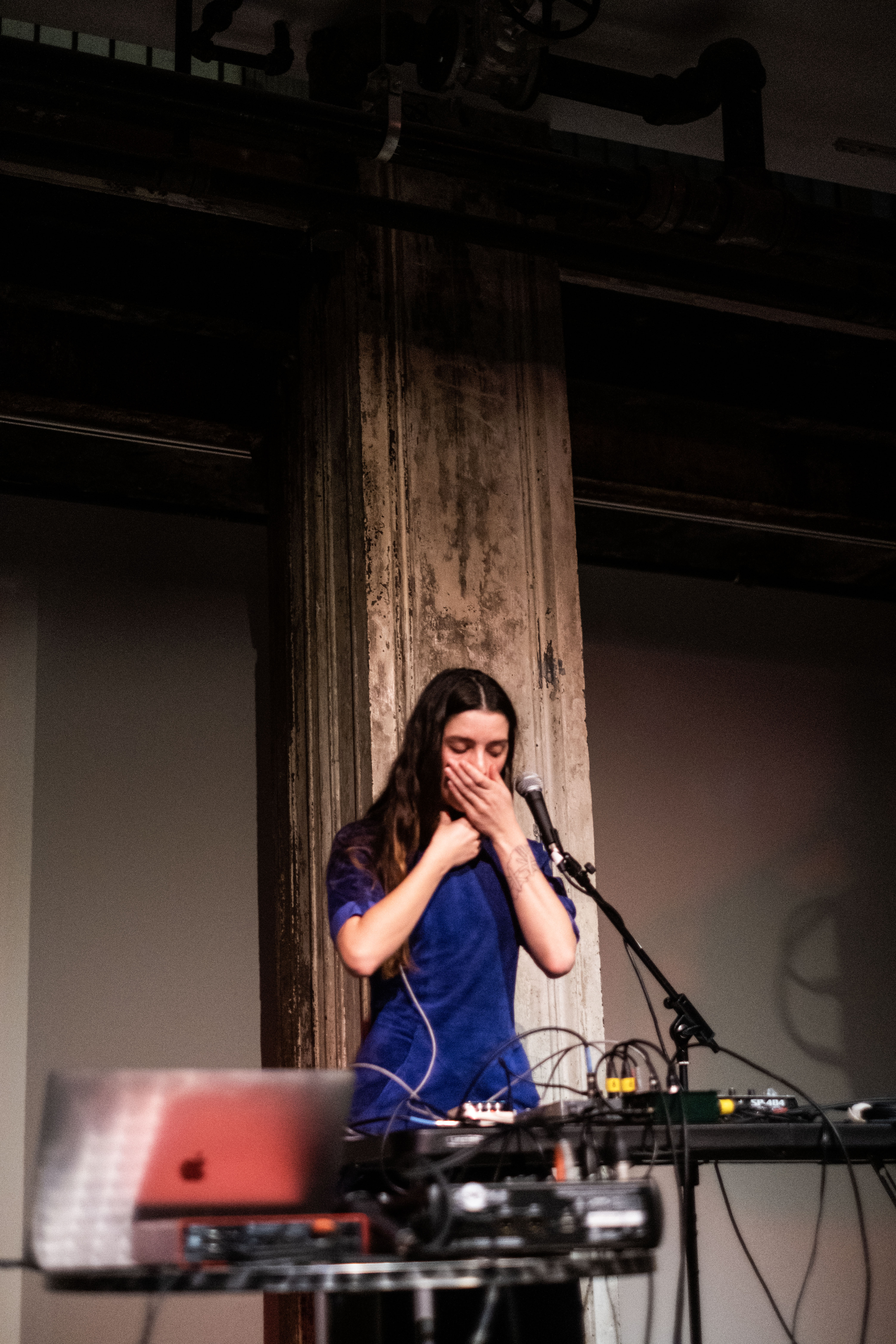 A figure stands behind a microphone and table with audio equipment. They hold one hand over their mouth and the other around their neck.