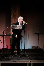A figure in a suit stands on stage, behind a music stand and speaks into a microphone.