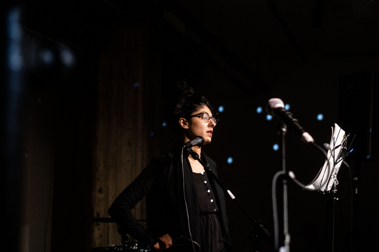 A figure is shown in profile performing on stage into a microphone, with a music stand placed in front of them.