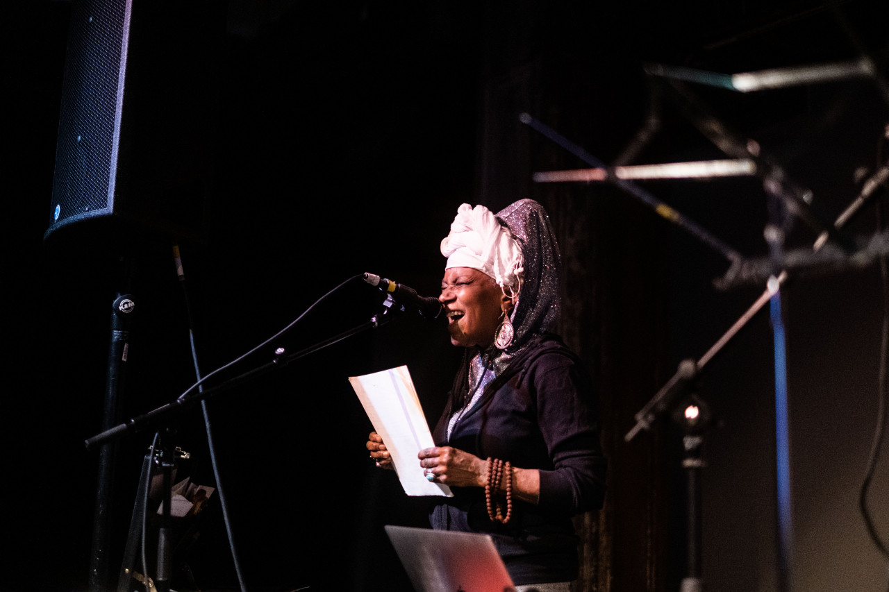 A figure performs on stage, speaking into a microphone and holding papers in their hands.