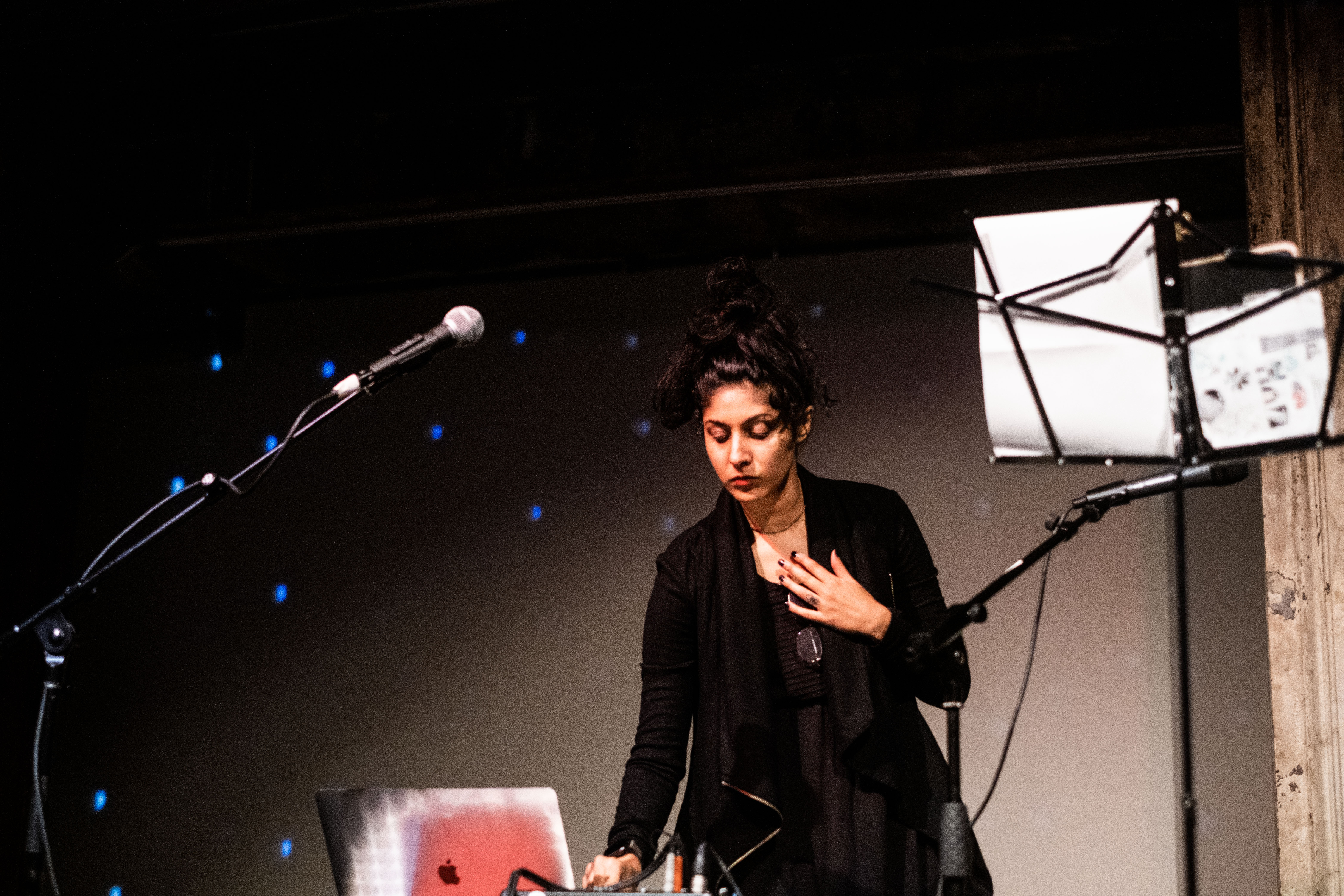 A figure stands on stage behind a table with a laptop, preparing to perform.