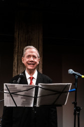 A figure smiles and stands behind a music stand and microphone.