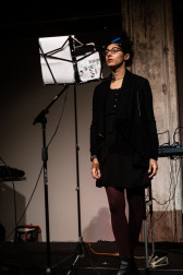 A figure performs on stage, standing straight and looking at material held on a music stand in front of them.