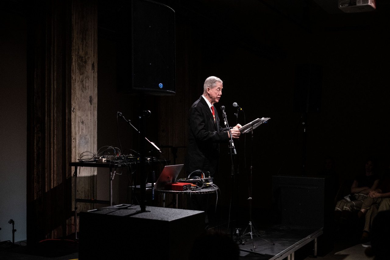 A figure in a suit performs on stage, reading into a microphone off sheets on a music stand in front of them.