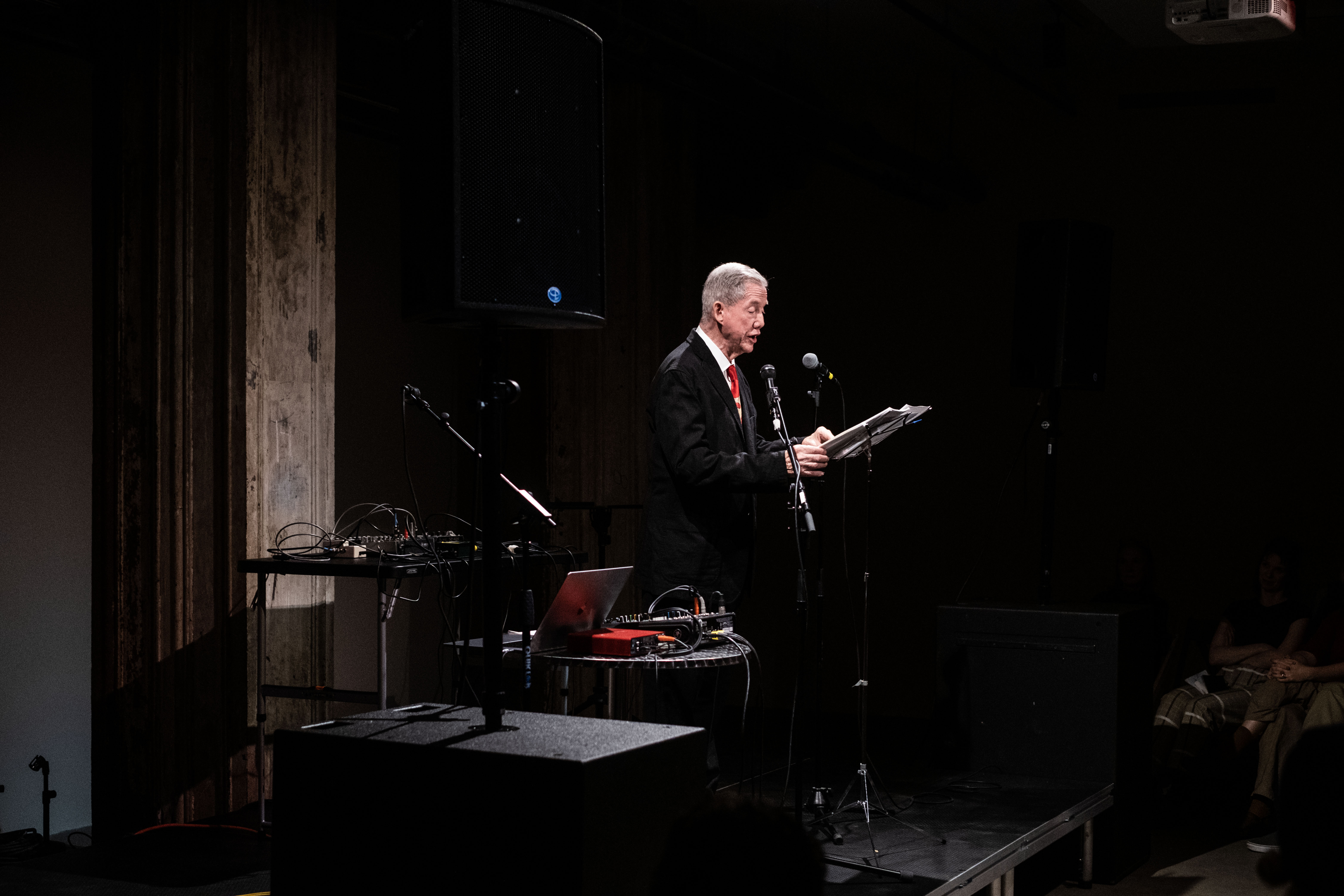 A figure in a suit performs on stage, reading into a microphone off sheets on a music stand in front of them.
