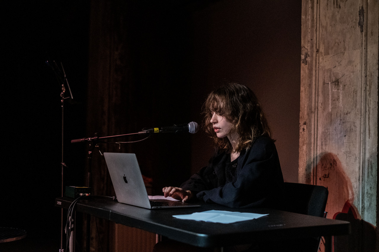 A figure is shown in profile speaking into a microphone, sitting behind a table and working on a laptop.