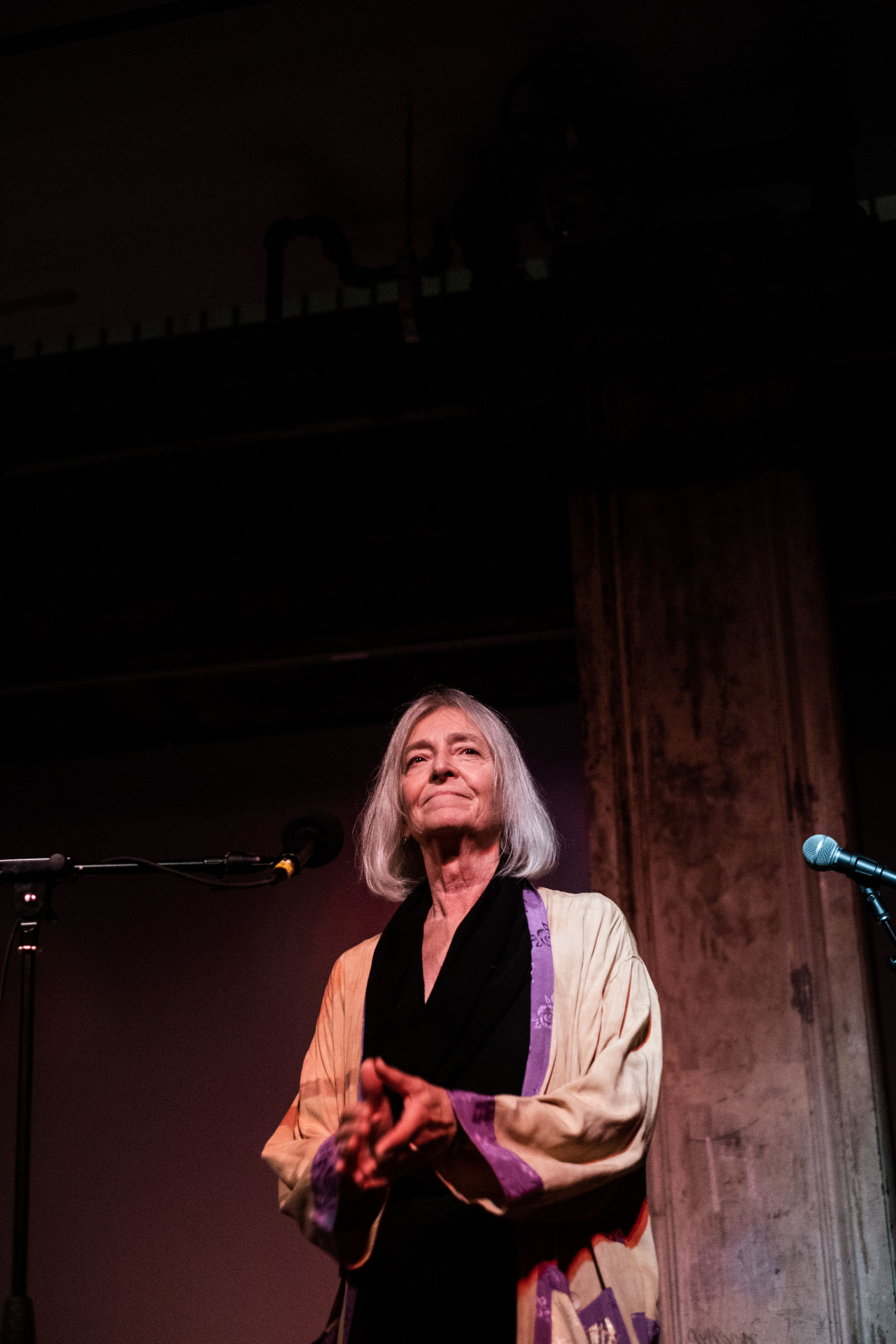 A figure stands behind a microphone, their hands placed together in front of them and looking off.