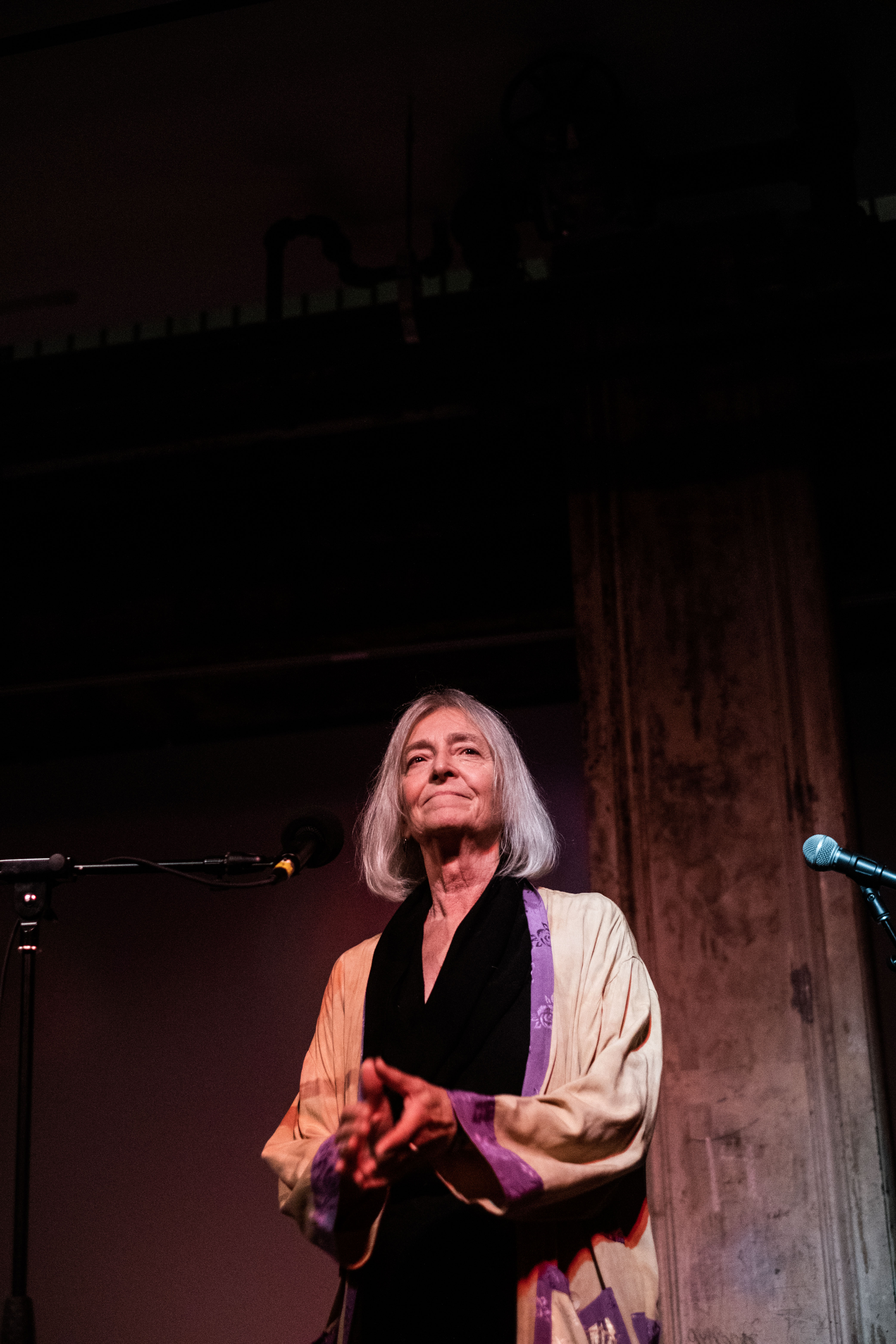 A figure stands behind a microphone, their hands placed together in front of them and looking off.