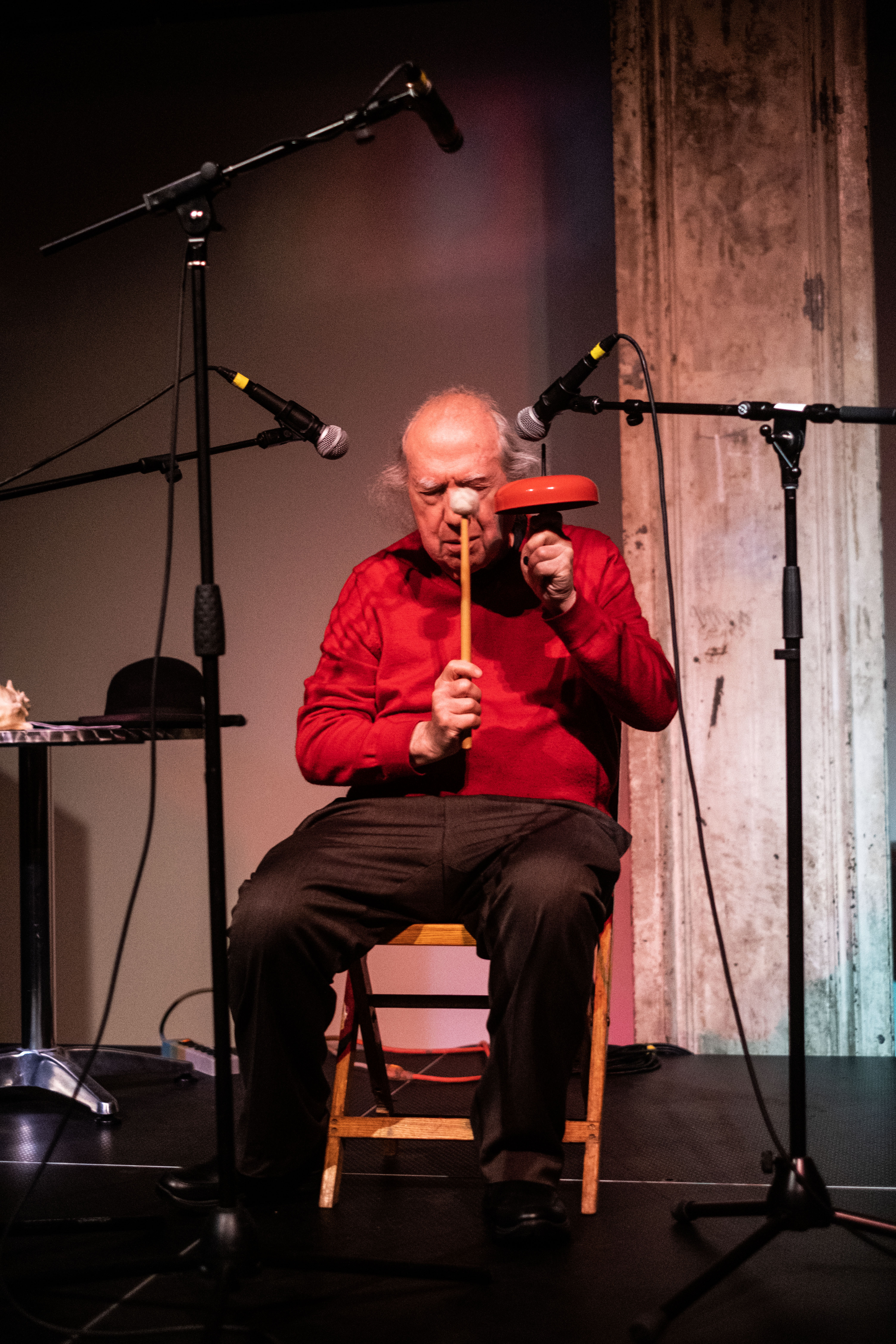 A figure in a red sweater sits in a chair on stage. Two microphones are positioned around them. They hold a wooden stick and a red bell up.