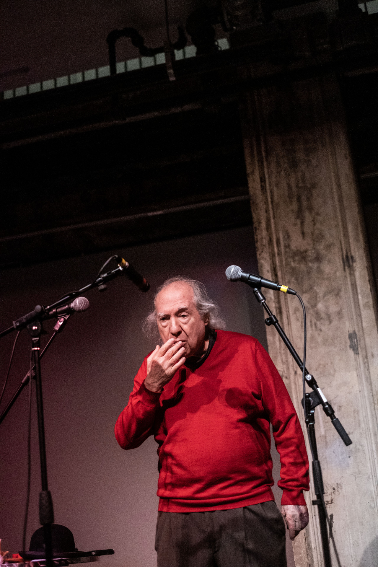 A figure in a red sweater stands on stage, behind three microphones placed around them, their left hand held up to their mouth.
